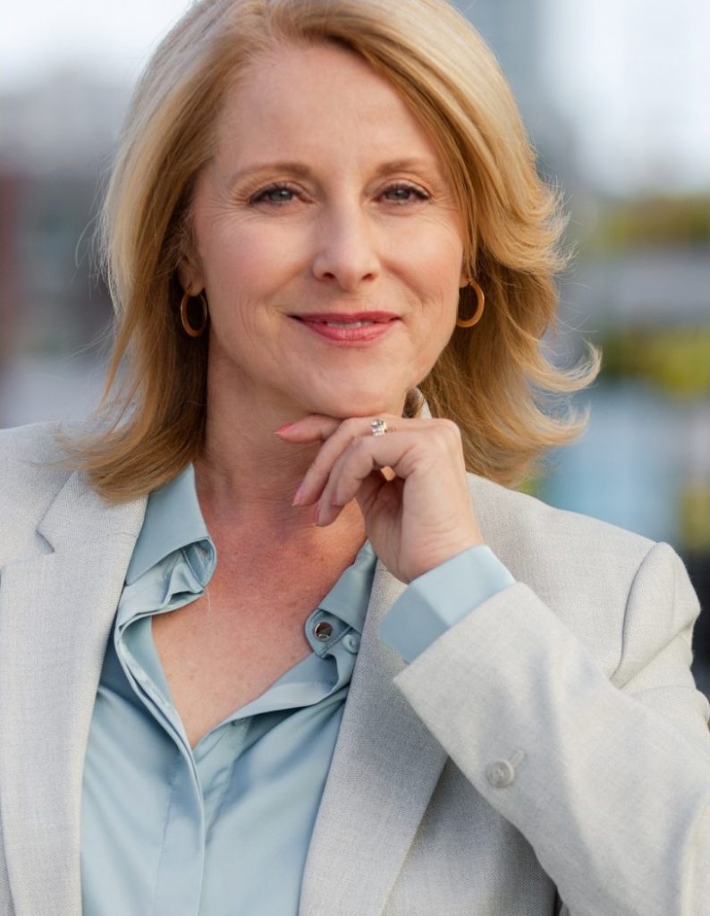 Professional woman smiling with hand on chin, wearing a pale blue blouse and light blazer.