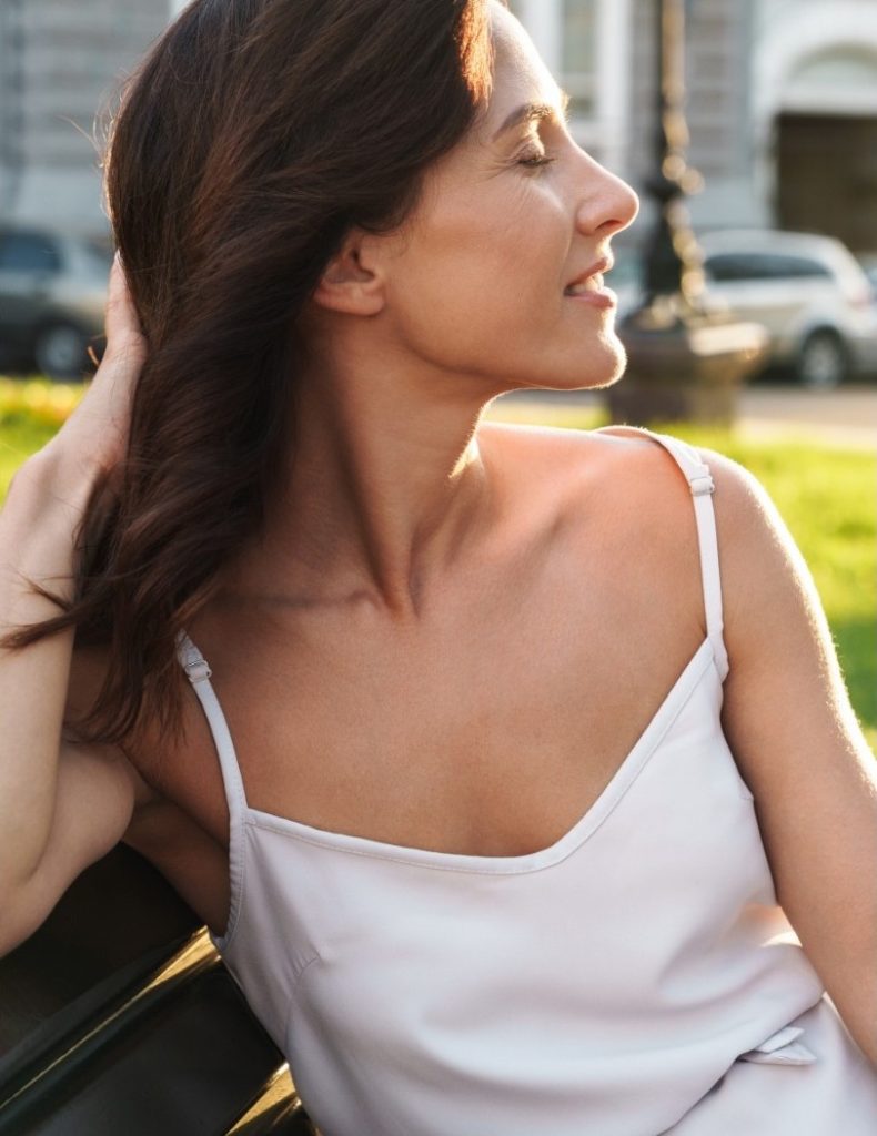 Woman in a white camisole sitting on a bench, smiling with eyes closed and sunlight on her face.