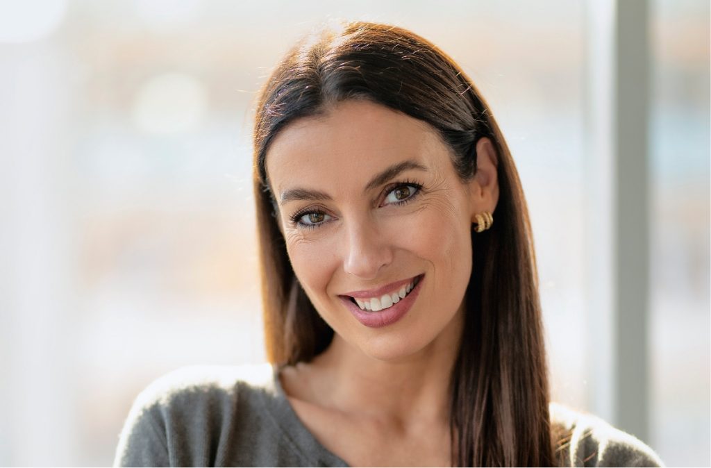 Woman with long straight brown hair smiling confidently in a white top and gray cardigan.