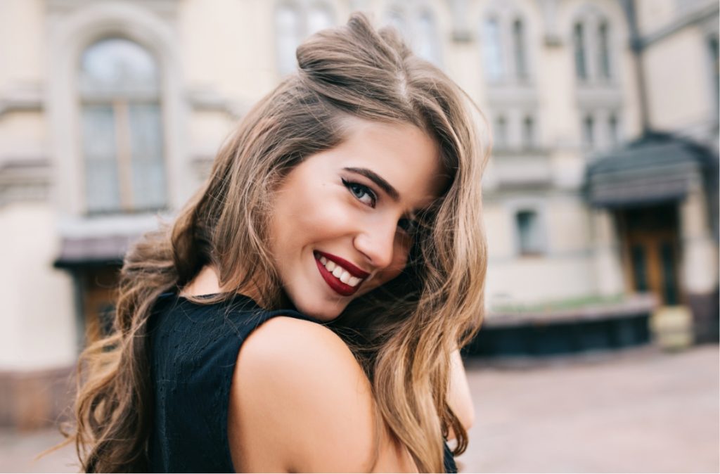 Young woman with winged eyeliner and red lipstick smiling over her shoulder in a black top.
