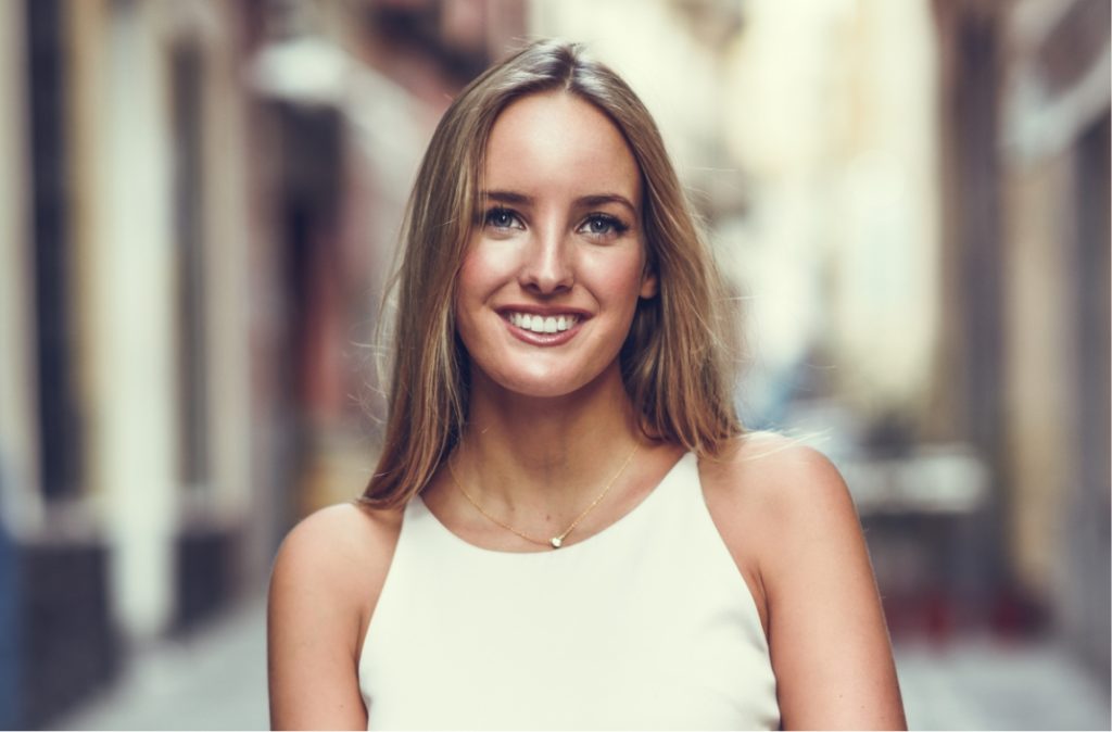 Smiling woman in a white sleeveless top standing outdoors with soft natural light.