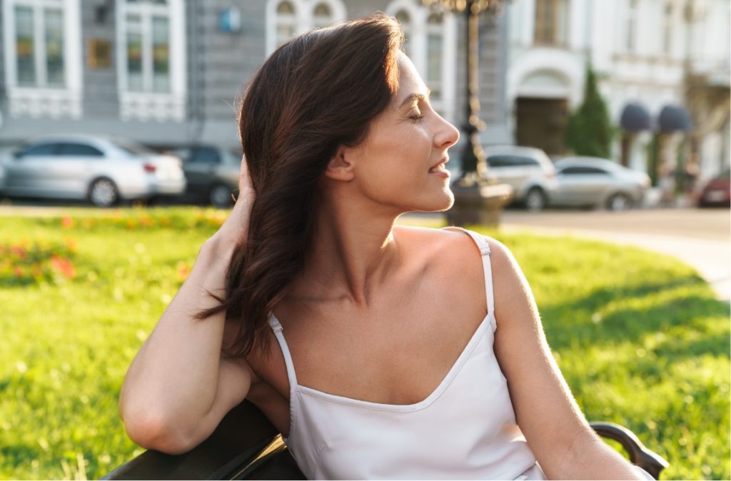 Woman in a white camisole sitting on a bench, smiling with eyes closed and sunlight on her face.