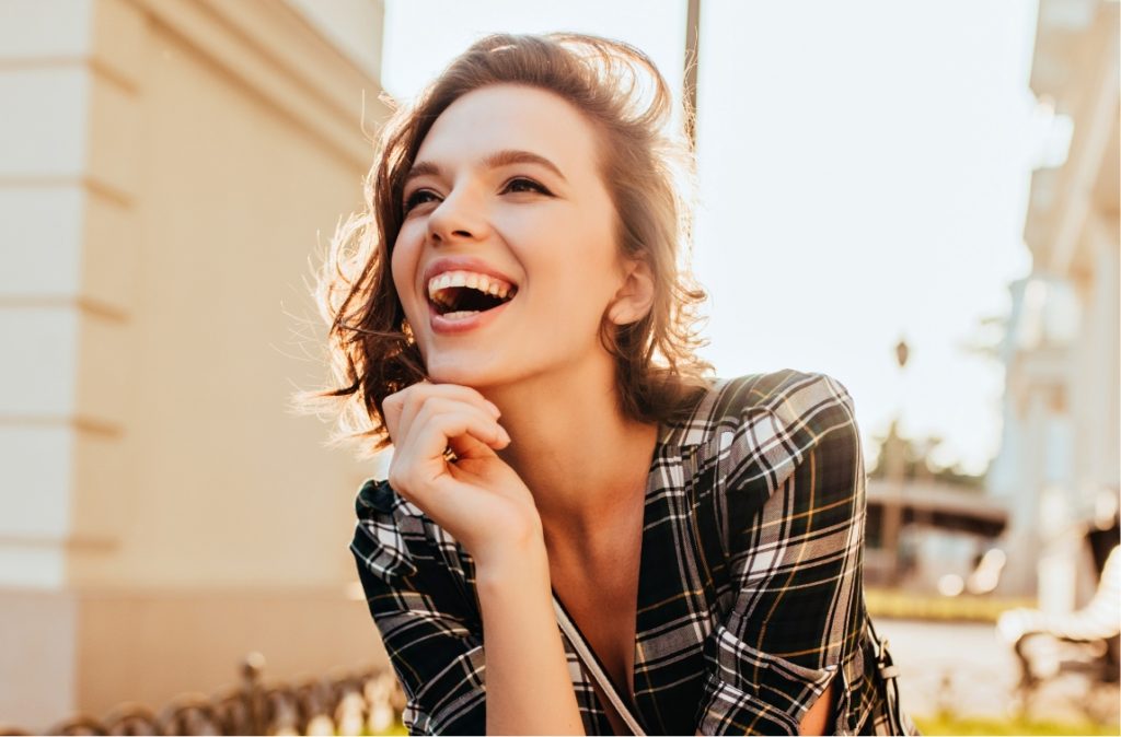 Woman in plaid dress laughing with hand on chin, lit by golden hour sunlight.